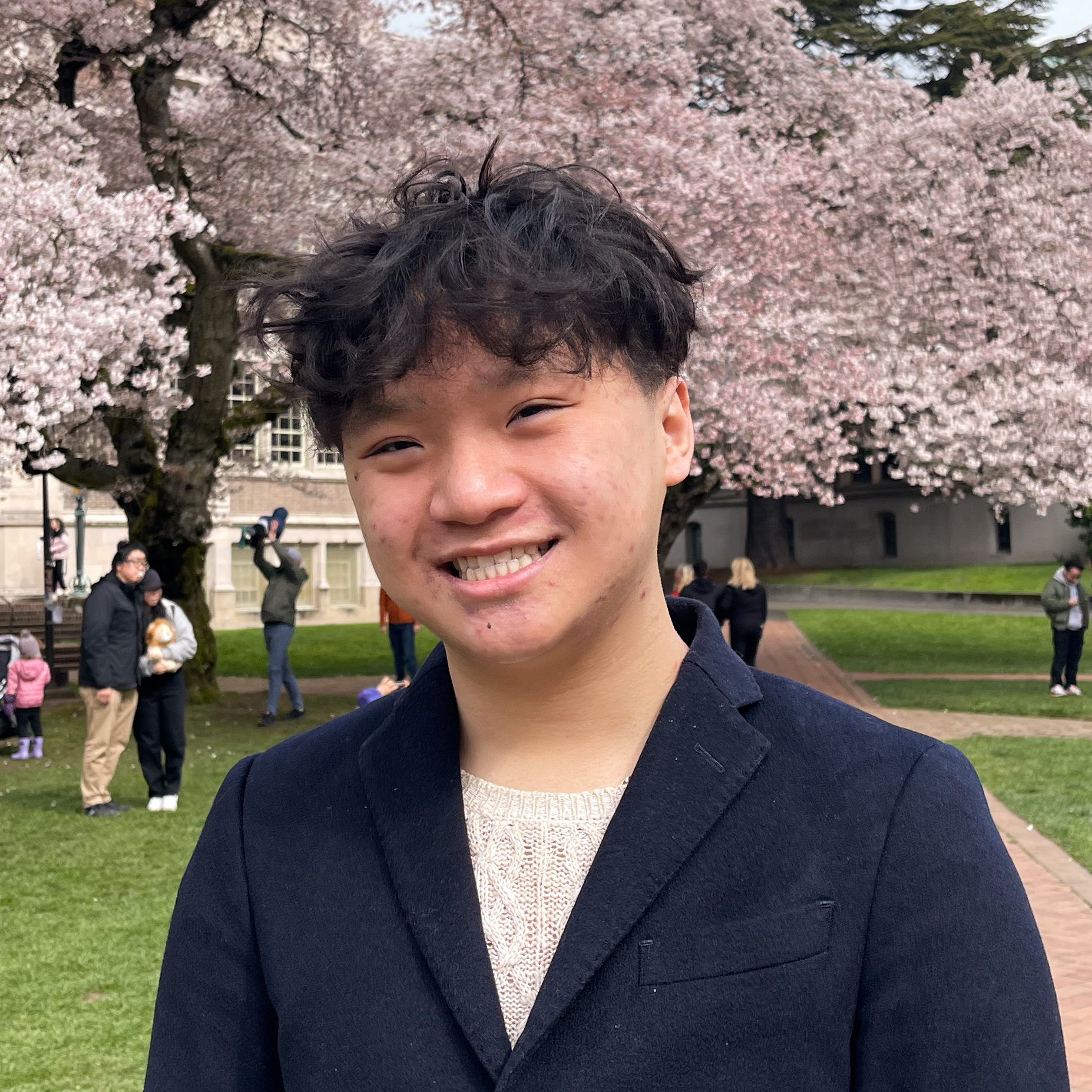 Ryan Le, Asian American male smiling with Curly-wavy hair with a black blazer and white sweater with pink cherry blossom trees in the background